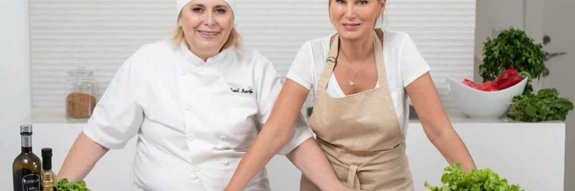 Two female chefs with variety of fresh ingredients in a kitchen setting, showcasing culinary preparation.
