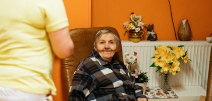 A warm scene of a senior woman sitting in an armchair at a retirement home with a caretaker nearby.