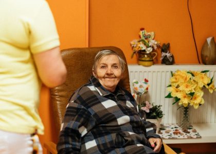 A warm scene of a senior woman sitting in an armchair at a retirement home with a caretaker nearby.